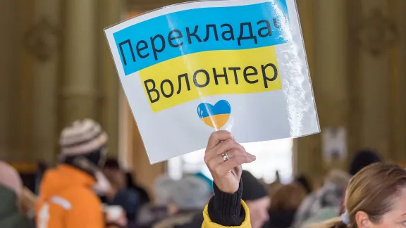 A woman holds up a sign offering practical support for newly arrived families from Ukraine, at Nyugati station in Budapest, Hungary. At the station, a range of civil society organisations and other volunteers offer support to incoming refugees, including support in arranging free accommodation, tickets for onward travel, as well as necessary items such as snacks and food, diapers for the children, clothes and basic medical supplies. Photo: LWF/Albin Hillert