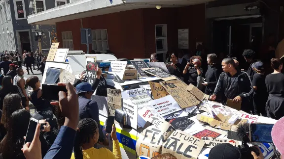 The anti-femicide demonstration in Cape Town, South Africa following the death of Uyinene Mrwetyana. Protestors cover a police vehicle with placards. Photo: Discott, via Wikimedia Commons (CC-BY-SA)